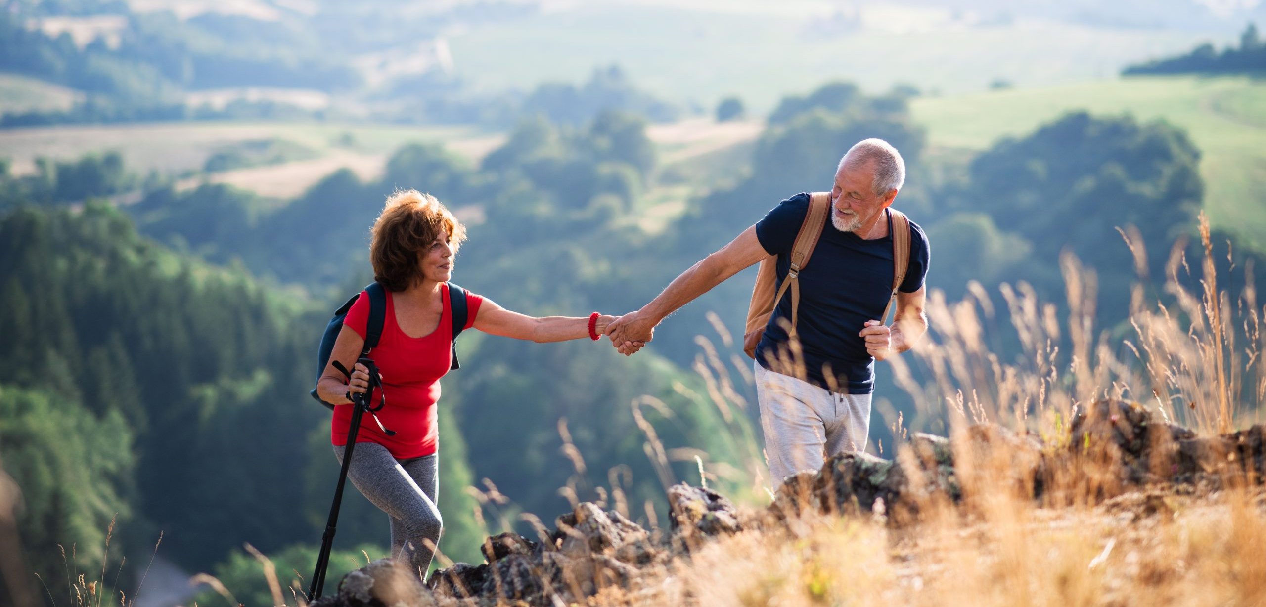 Senior tourist couple with backpacks hiking in nature, holding hands.